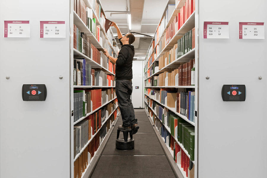 Person standing on a step stool, reaching for books on high shelves in compact shelving.